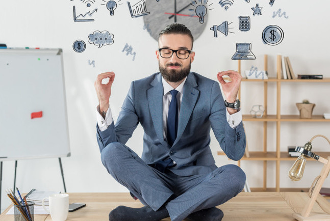 A young man in a professional suit sits cross-legged in an office with his eyes closed and his hands in a meditative pose while he smiles and 2 dimensional ideas float around his head