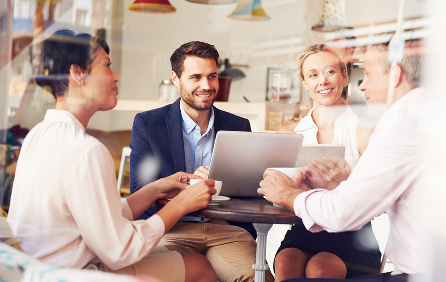 4 men and women sit around a table discussing business over a laptop all warmly smiling at each other