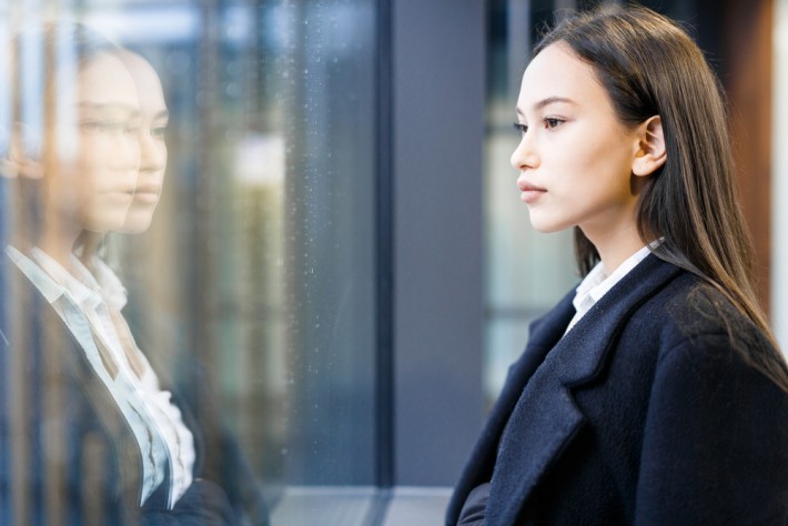 A young Asian woman in a suit jacket stares out of a rainy window with a blank expression on her face