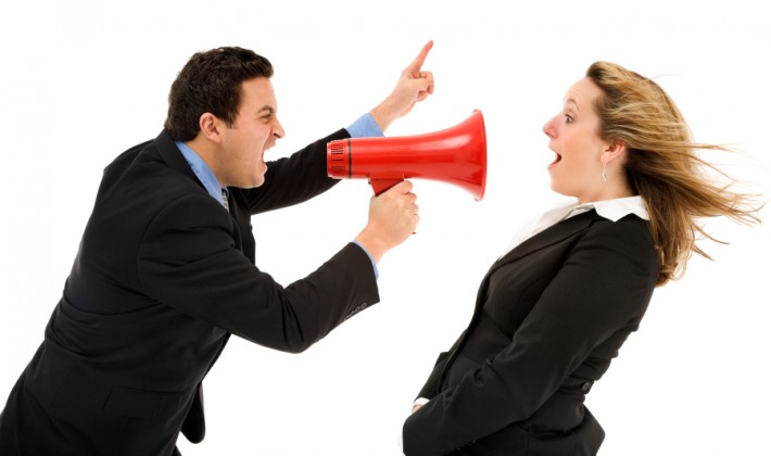 A man in a suit yells through a megaphone angrily at a young woman in front of him also wearing a similar suit looking surprised
