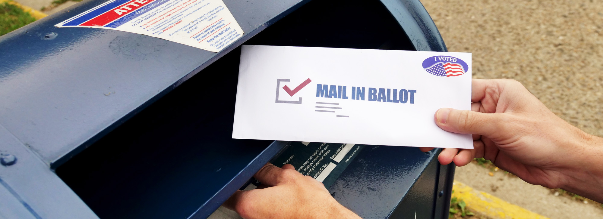 Someone drops off an envelope into a mailbox that reads "Mail In Ballot"