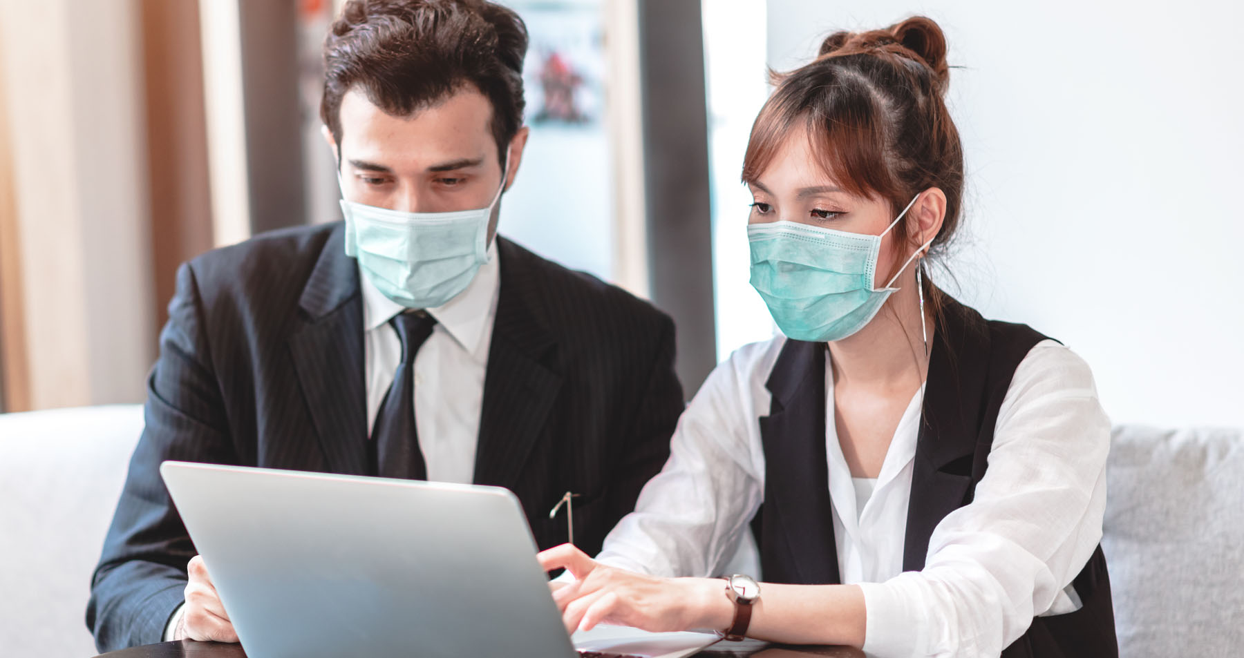 A young man and woman dressed in business attire with PPE masks on sit next to each other reviewing something on an open laptop in front of them