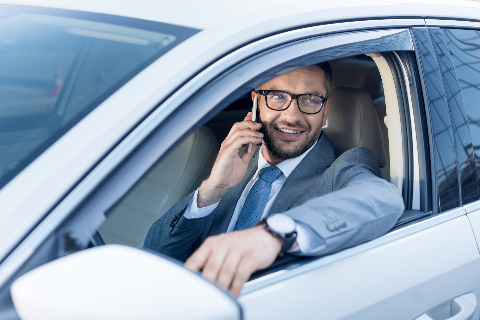 A man in business attire sits in his car with the window rolled down looking outward as he speaks to someone on his phone smiling