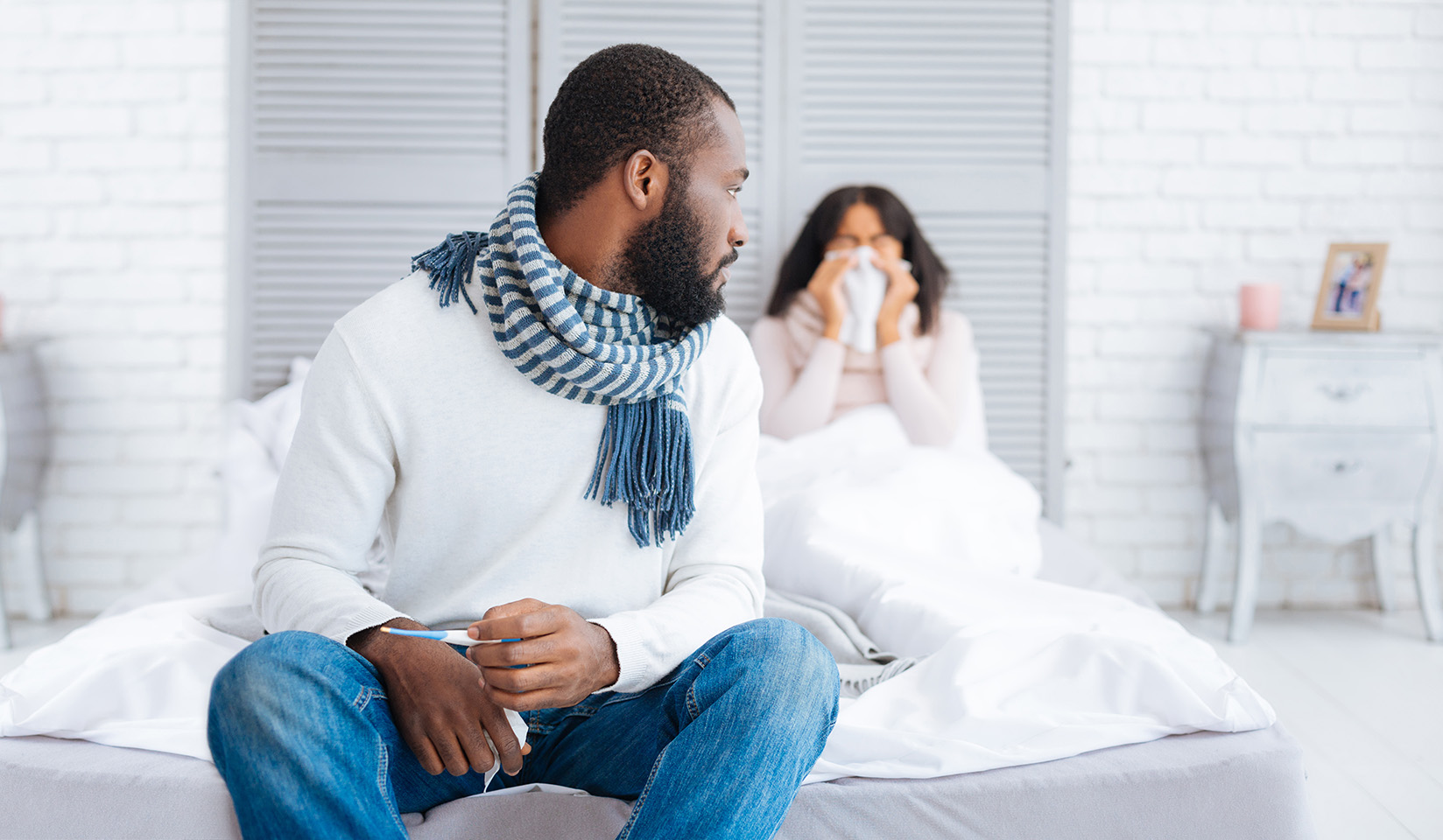 A black man sits on the foot of a bed in the foreground turning around to look at a woman sitting behind him under the blanket blowing her nose into a tissue indicating she is sick