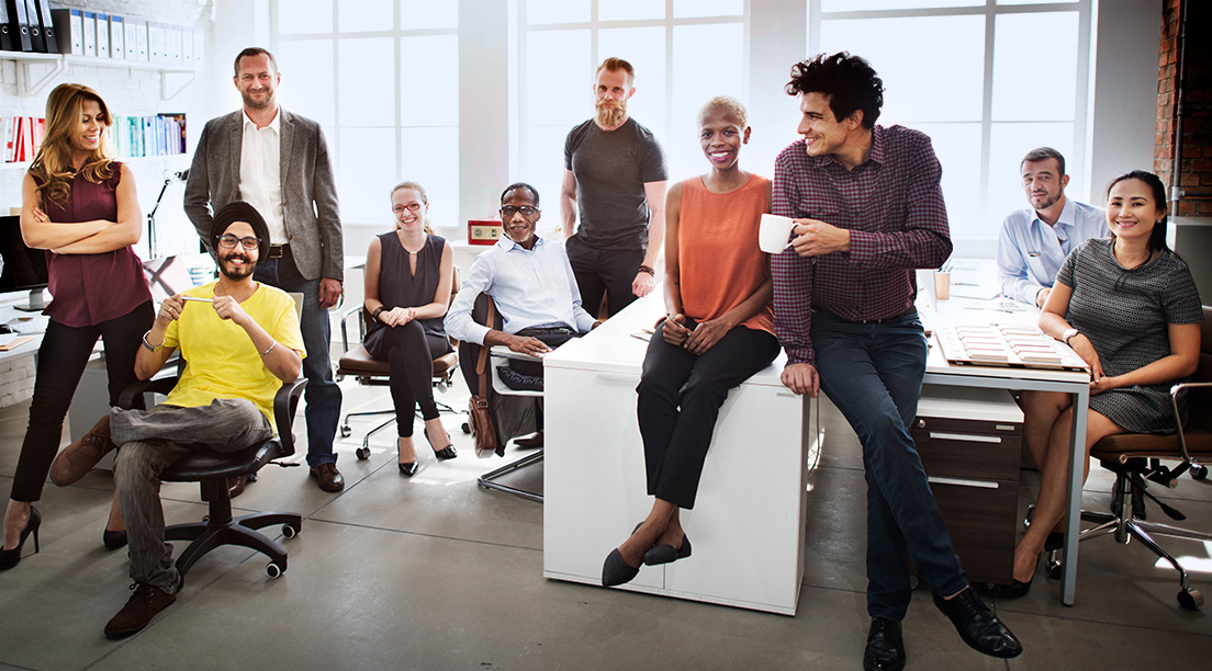 10 diverse men and women sit in and stand in an office smiling and either engaging with one another or looking directly at the camera