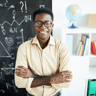 A young black man stands in front of a chalkboard with his arms crossed while smiling warmly.
