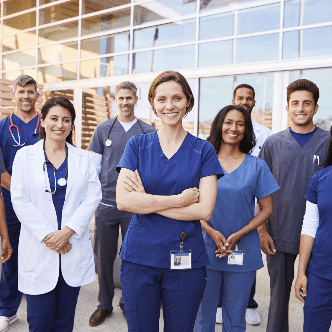 A team of men and women wearing scrubs and stethoscopes stand in front of a hospital smiling.