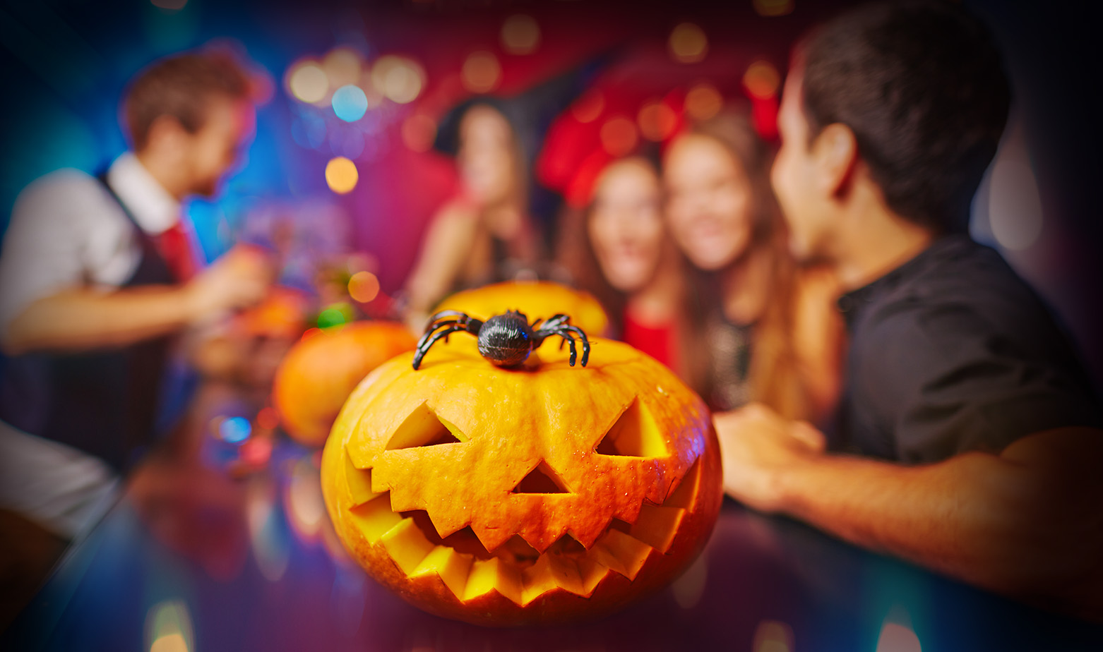 A Jack-O-Lantern sits on a party table with a plastic spider on top of it seemingly at a work Halloween party