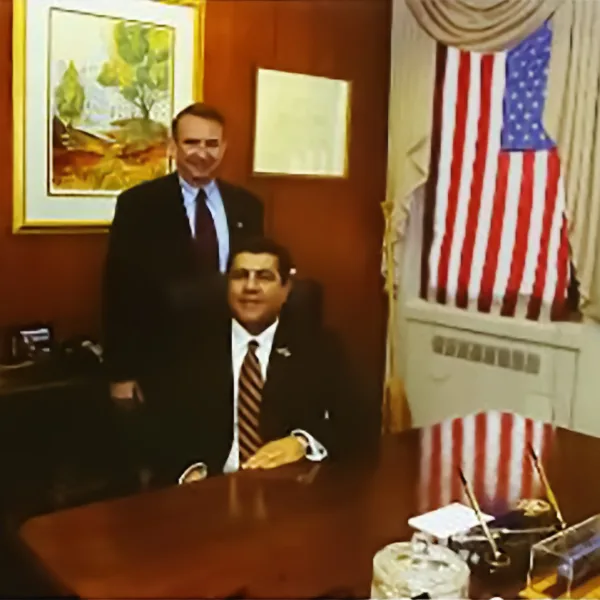 2 men, one standing and one sitting, in an office dressed in suits behind a desk